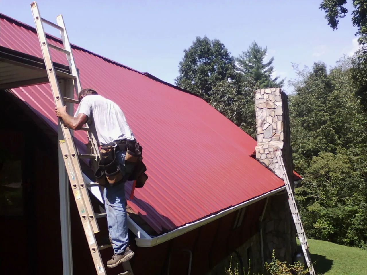 Roofer on a ladder beside a red metal roof and stone chimney — Soffit & Fascia Repair by Roofing Brothers in West Bend